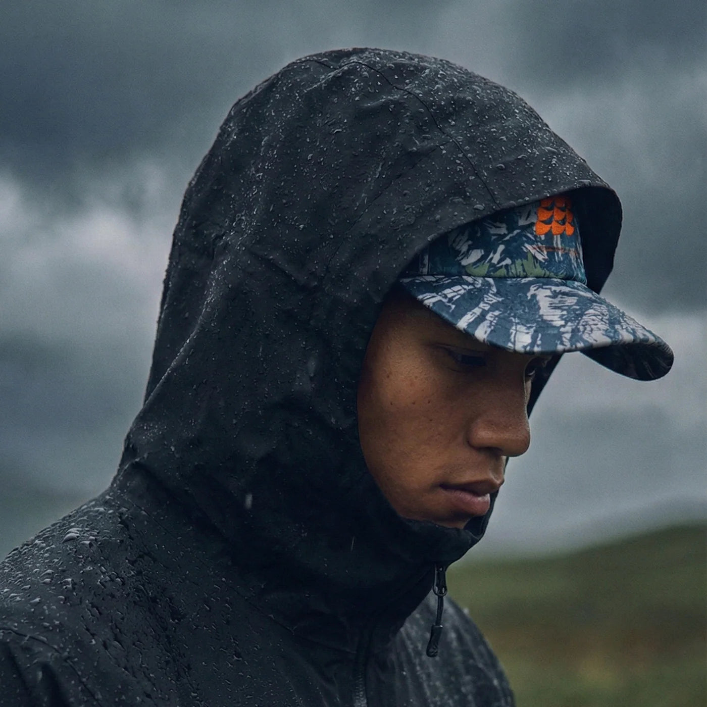 Person wearing a dark raincoat and camouflage cap in a rainy outdoor setting